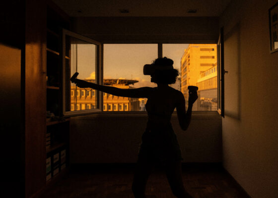 A woman standing in what seems to be here flat wearing a virtual reality device. In the background you can see through a window and get a glimpse of neighbour buildings.