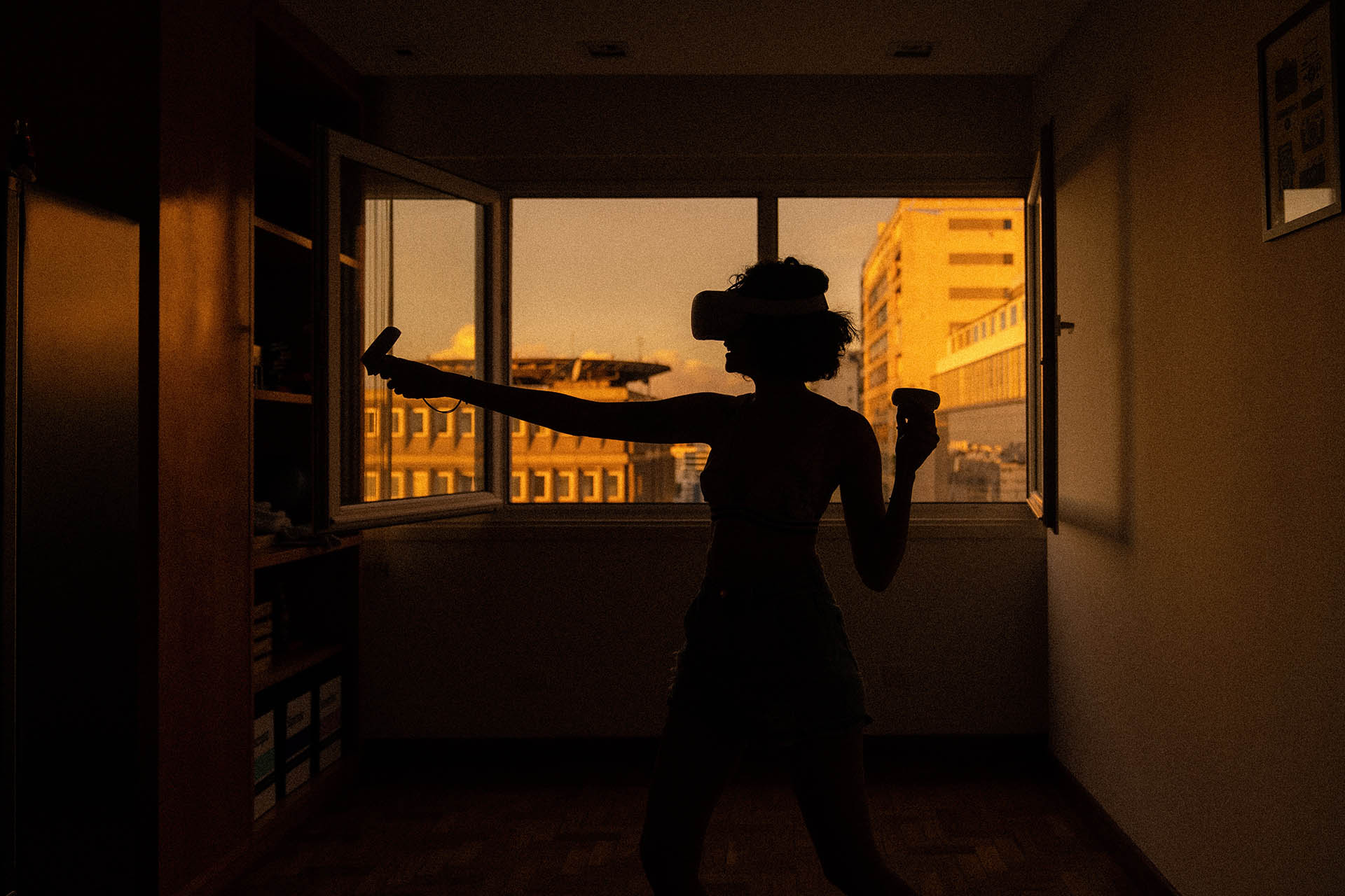 A woman standing in what seems to be here flat wearing a virtual reality device. In the background you can see through a window and get a glimpse of neighbour buildings.