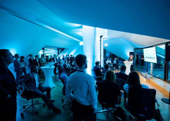 Blue top floor of a building with sloping roofs on the left and right side. At least 40 people are visible. The light is blue and rather dark. On the right side you can see a speaker holding a keynote in front of a screen, on the right side the audience is listening.