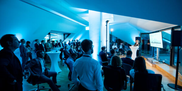 Blue top floor of a building with sloping roofs on the left and right side. At least 40 people are visible. The light is blue and rather dark. On the right side you can see a speaker holding a keynote in front of a screen, on the right side the audience is listening.