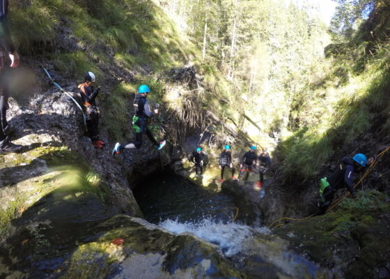 An accilium employee is jumping from a cliff inside a canyon into a big natural pool. Several other accilium employees are standing aside and applaude him.