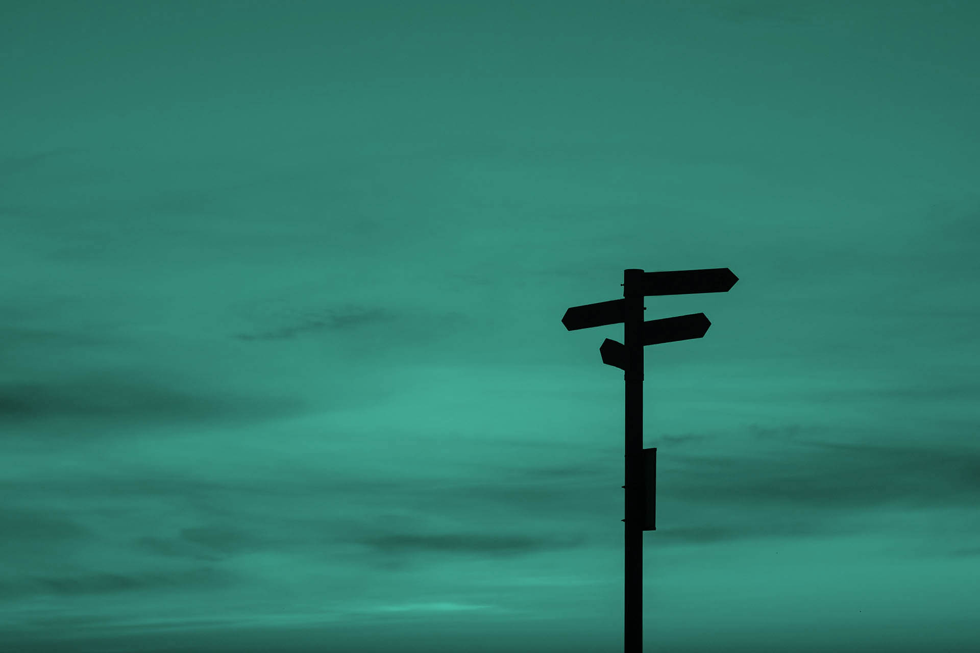 Directional signs, which can only be recognised as black outlines due to the contrast and incidence of light, stand in front of a bluish-red horizon with individual clouds.