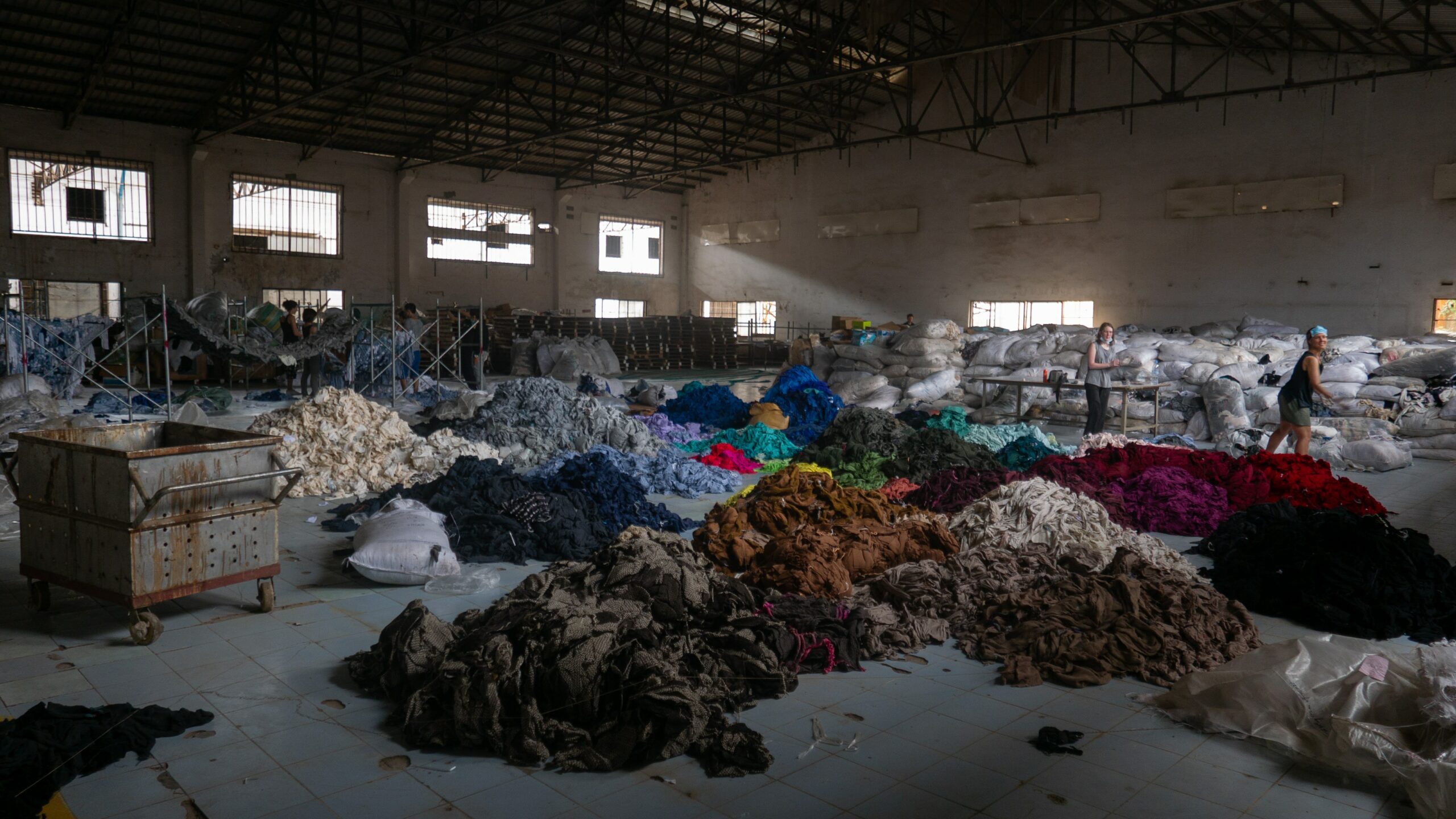 A huge industrial hall with many piles of textile garbage in different colours on the floor. Two women carrying some textiles from the center to the right.